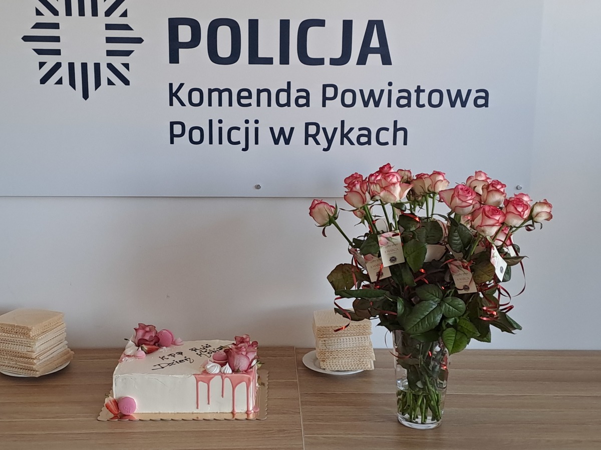 Logo Policji Polskiej z napisem Komenda Powiatowa Policji w Rykach oraz kwiaty w wazonie i tort z okazji Dnia Kobiet