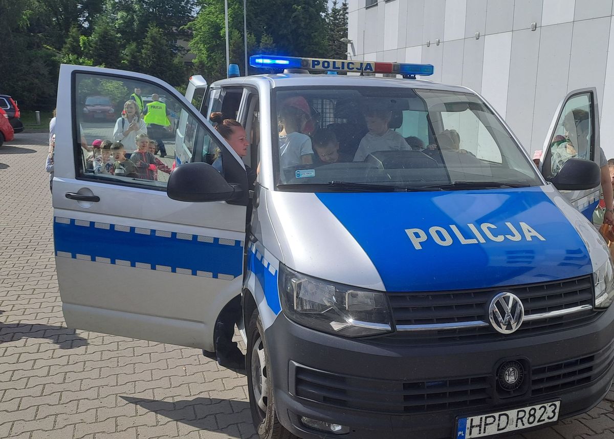 Zdjęcie przedstawia radiowóz typu bus oznakowany koloru srebrnego który jest zwiedzany przez przedszkolaków na placu komendy policji w rykach
