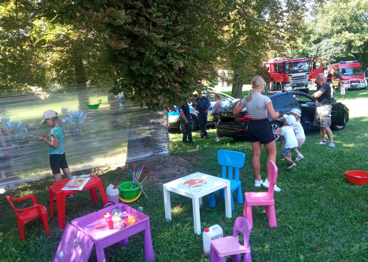 Wiodczny plener pikniku w pierwszym tle stoliki i krzesła dla dzieci folia do malowania a w tle pojazdy policji i strażacy. Wokół bawią się dzieci młodzież i rodzice