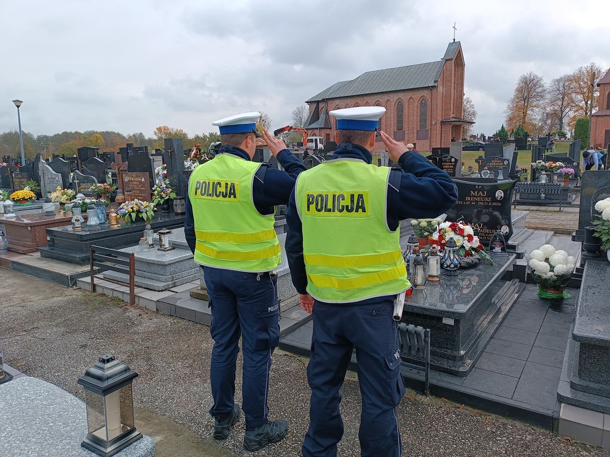 Dwóch policjantów stojących tyłem oddaje hołd zmarłemu koledze stojąc przed jego nagrobkiem na cmentarzu