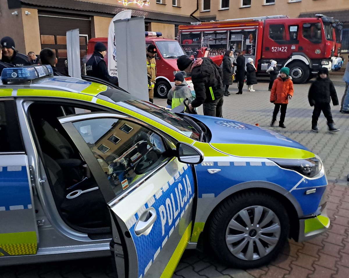 Stoisko Komendy Powiatowej Policji w Rykach na finale w WOSP w Rykach na placu OSP przy kinie Renesans. Widoczne pojazdy służbowe radiowozy oznakowane i nieoznakowane, osoby odwiedzające i sprzęt policyjny i straży pożarnej.