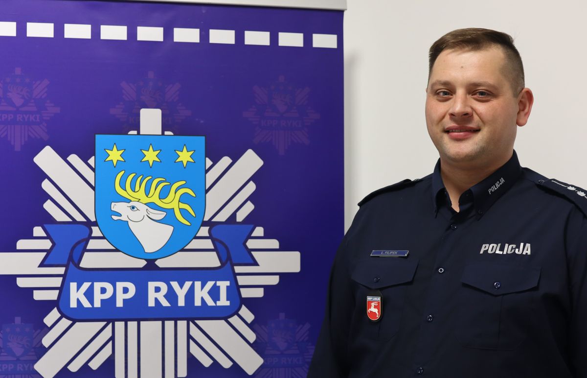 oficer prasowy ||aspirant Łukasz Filipek obok logo Komendy Powiatowej Policji w Rykach