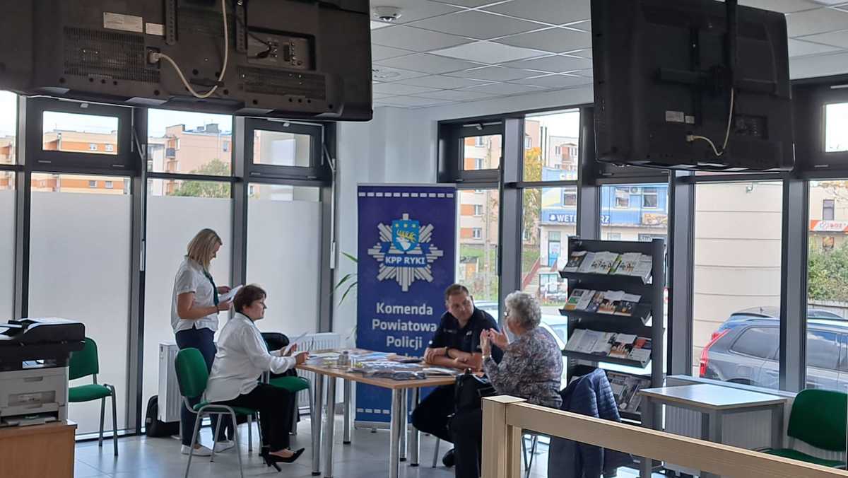 Stoisko Policji z Rykach w Zakładzie Ubezpieczeń Społecznych w Rykach na którym jest policjant oraz pracownicy ZUS-u i seniorka