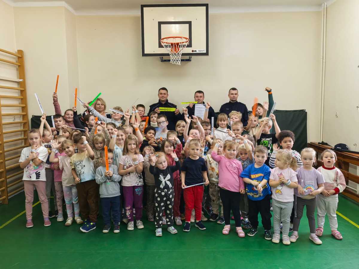 Zdjęcie grupy uczniów pokazujących odblaski oraz policjantów i nauczycieli na sali gimnastcznej