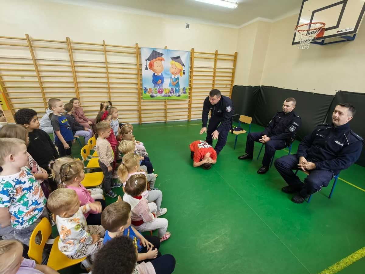 Policjant wraz z jednym z uczniów prezentuje pozycje tak zwanego żółwia grupie dzieci