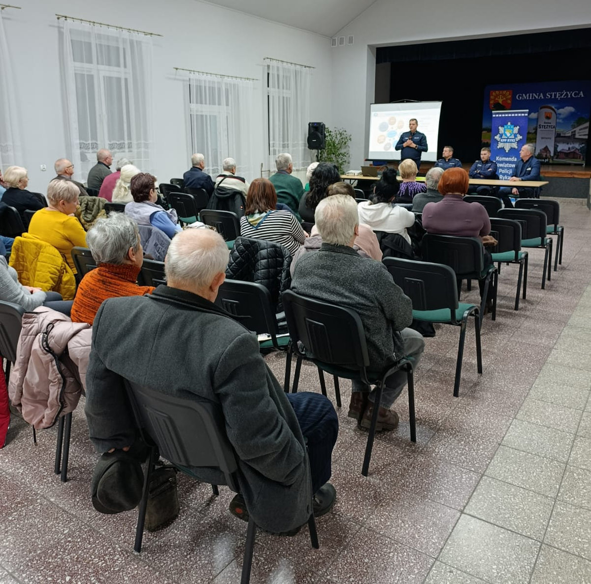 Uczestnicy debaty na sali konferencyjnej GOK w Stężycy siedza na krzesłach policjant przemawia