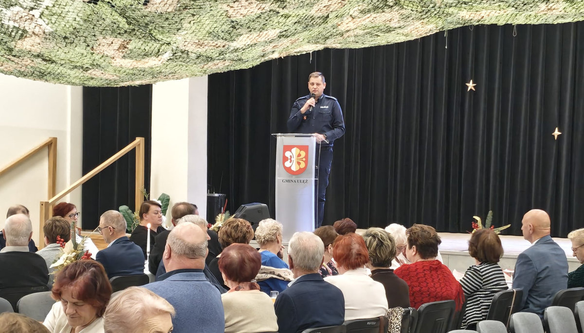 Policjant przemawia przed grupa uczestników spotkania w sali w GOK w Ułężu