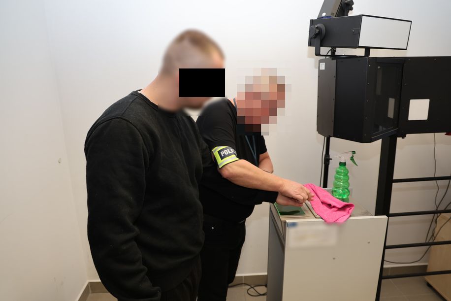 Policjant technik kryminalistyki pobiera odbitki placów urządzeniem elektronicznym od zatrzymanego mężczyzny