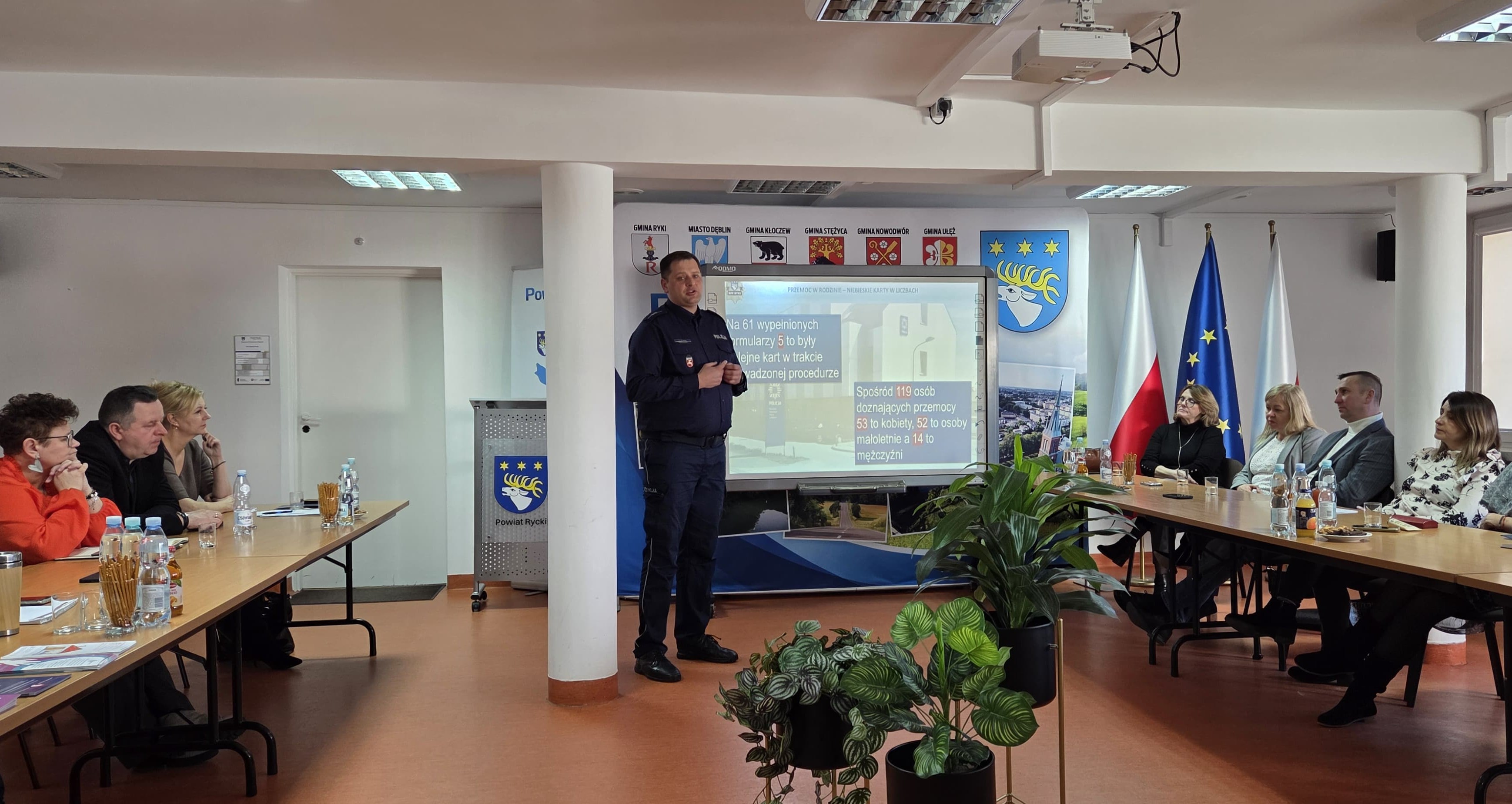 Policjant podczas prelekcji obok przy stołach siedzą uczestnicy w tle wyświetlana jest prezentacja