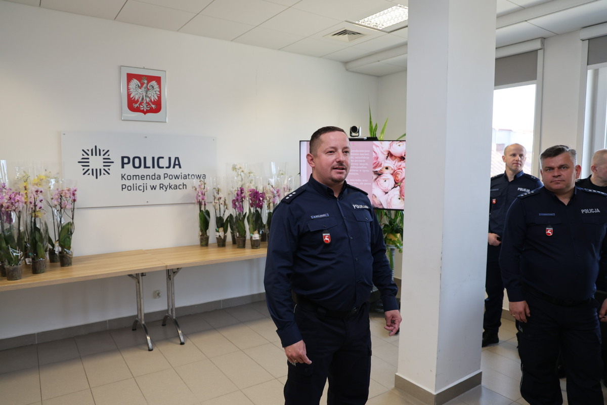 I Zastępca Komendanta Powiatowego Policji w Rykach składa życzenia na sali odpraw w tle funkcjonariusze i kwiaty