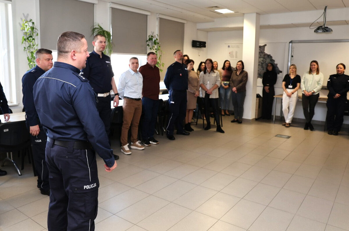 I Zastępca Komendanta Powiatowego Policji w Rykach składa życzenia na sali odpraw w tle funkcjonariusze i kwiaty