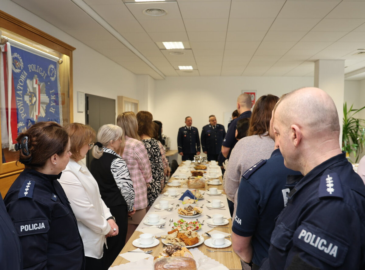 funkcjonariusze i pracowenicy ryckiej komendy na spotkaniu wigilijnym stoją z obu stron przy nakrytym stole z  potrawami w tle widoczna kadra kierownicza