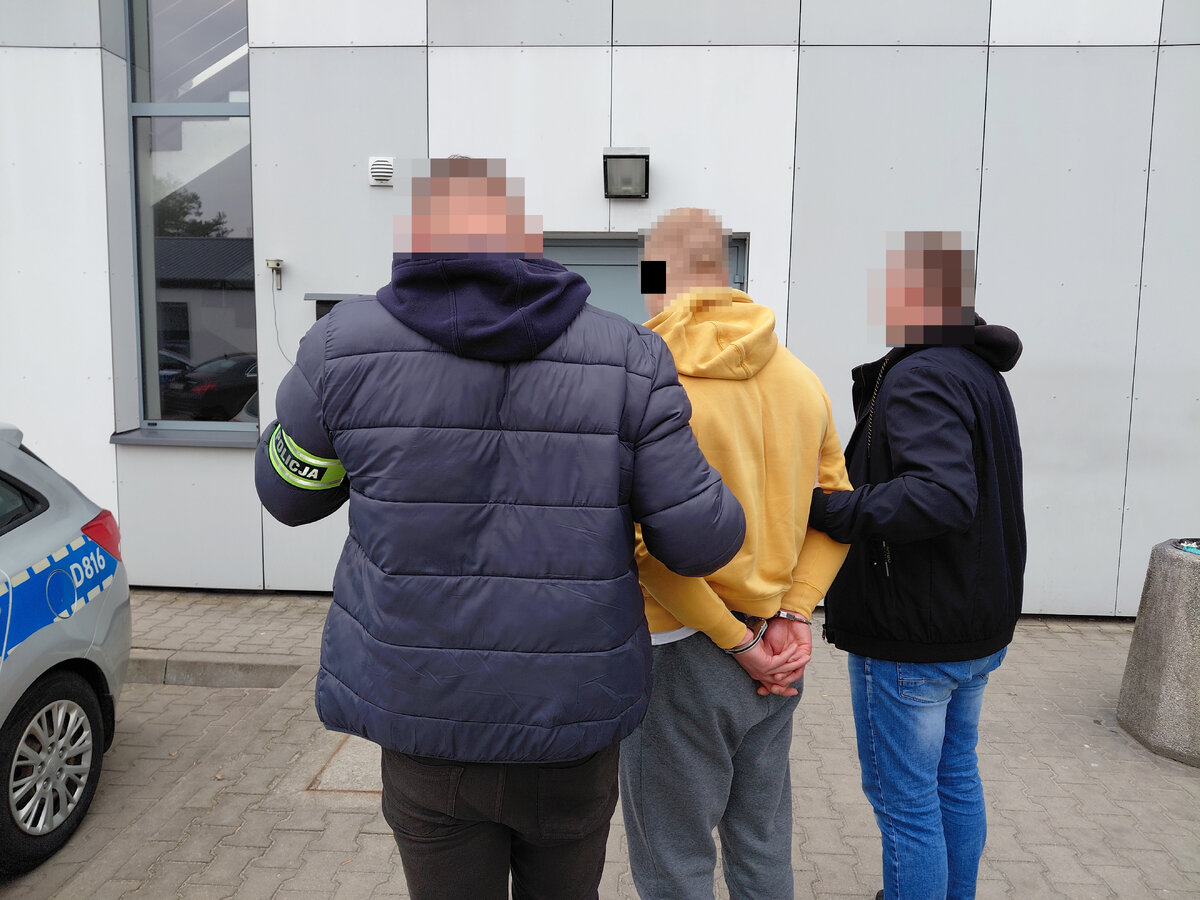 Dwóch policjantów prowadzi zatrzymanego mężczyznę z kajdankami założonymi na ręce z tyłu. W tle budynek i radiowóz policyjny. 