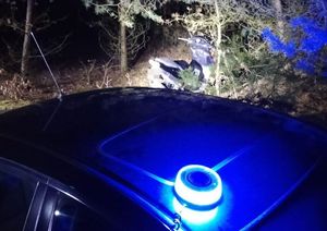 Na zdjęciu widać nieoznakowany radiowóz Policji z włączonym sygnałem świetlnym koloru niebieskiego oraz oparty o drzewo motorower.