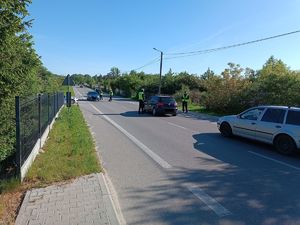 Zdjęcie przedstawia funkcjonariuszy Wydziału Ruchu Drogowego KPP w Rykach którzy dokonują badania trzeźwości kierujących na drodze powiatowej o nawierzchni asfaltowej. Funkcjonariusze są umundurowani i mają żółte kamizelki odblaskowe.