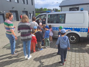 Zdjęcie przedstawia radiowóz typu bus oznakowany koloru srebrnego który jest zwiedzany przez przedszkolaków na placu komendy policji w rykach
