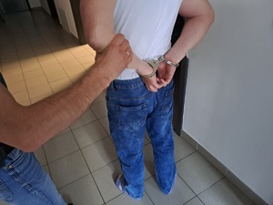 Na zdjęciu jest widoczny mężczyzna stojący tyłem ubrany w białą koszulkę oraz niebieskie spodnie na ręce trzymane z tyłu ma złożone kajdanki i za lewą rękę jest trzymany przez policjanta służby kryminalnej