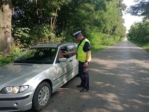 Zdjęcie przedstawia umundurowanego funkcjonariusza pionu ruchu drogowego z założoną kamizelka odblaskową koloru żółtego podczas kontroli drogowej stojącego obok samochodu osobowego.