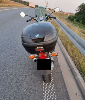 Zdjęcie przedstawia motocykl marki BMW stojący tyłem. Motocykl ma zamontowany kufer koloru czarnego. Pojazd stoi na drodze asfaltowej z lewej strony są bariery energochłonne w oddali widać inne pojazdy