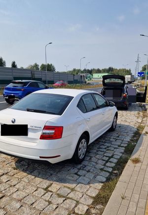 Jest to zdjęcie przedstawiające samochód osobowy koloru białego marki Skoda a w tle są widoczne inne pojazdy. Auto stoi na parkingu.