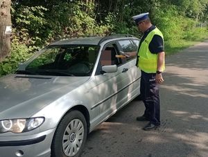 Zdjęcie przedstawia funkcjonariusza umundurowanego pionu ruchu drogowego stojącego obok pojazdu koloru srebrnego marki BMW . Policjant jest w trakcie badania stanu trzeźwości