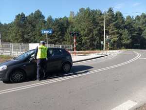 Na zdjęciu widoczny jest policjant ruchu drogowego umundurowany stojący obok pojazdu kontrolowanego jest samochód osobowy koloru zielonego. Kontrola jest wykonywana na drodze w pobliżu przejazdu kolejowego. Policjant stoi odwrócony plecami.