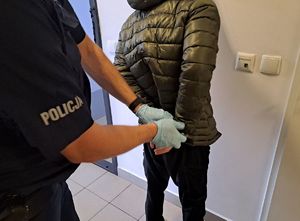 Zdjęcie jest zrobione na korytarzu pomieszczania dla osób zatrzymanych . Umundurowany policjant zakłada kajdanki na ręce z tyłu mężczyźnie ubranemu w zielona kurtkę i czarne spodnie. Policjant stoi bokiem na zdjęciu a mężczyzna odwrócony plecami. Zdjęcie przedstawia tułów obydwu mężczyzn - bez ich twarzy.