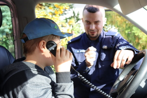 Zdjęcie przedstawia dziecko siedzące w radiowozie które trzyma pilot od sterowania sygnałami uprzywilejowani. koloru szarego. Dziecko zasłoniło nim swoją twarz ubrane jest w czapkę koloru niebieskiego. w tle widoczny jest policjant umundurowany stojący oko radiowozu.