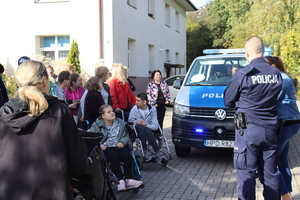 Zdjęcie przedstawia wychowanków ośrodka i ich wychowawców stojących na placu obok radiowozu. Przed nimi stoi policjant umundurowany. Grupa kilkunastu osób stoi w tym dwoje dzieci siedzi na wózku inwalidzkim.