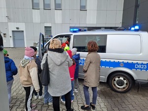 Zdjęcie przedstawia grupę dzieci ze szkoły podstawowej nr 2 w Dęblinie ubrane w różne kolorowe ubrania, Dzieci zwiedzają obiekt Komendy Powiatowej Policji w Rykach i zapoznają się ze sprzętem wykorzystywanym w słuzbie.