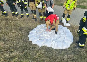 Zdjęcie przedstawia worki z pisakiem koloru białego ułożone w celu uszczelnienia przecieku wału. Wokół stoją strażacy.