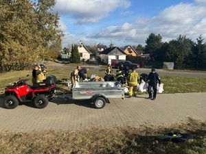 Na zdjęciu widoczna jest grupa strażaków oraz policjant którzy nasypują worki z pisakiem które ładują na przyczepkę podczepiona do quada