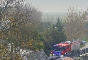 Widoczny jest pojazd wóz bojowy straży pożarnej obok strażak. Pojazd stoi przy ulicy gdzie są budynki mieszkalne.