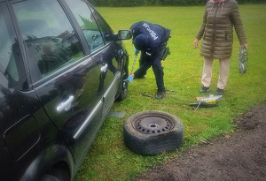 Policjant w trakcie zmiany koła w samochodzie obok stoi kobieta - twarz zamaskowana a obok policjanta  stoi auto koloru czarnego.