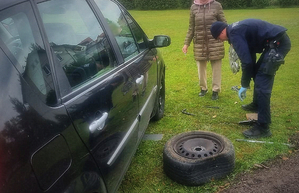 Policjant w trakcie zmiany koła w samochodzie obok stoi kobieta obok policjanta  stoi auto koloru czarnego.