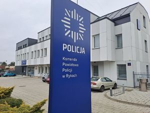 Zdjęcie przedstawia budynek Komendy Powiatowej Policji w Rykach oraz parking przed budynkiem w raz z banerem z logiem policji i nazwa komendy. Budynek ma elewację koloru biało szarego a baner jest niebieski z białymi napisami.