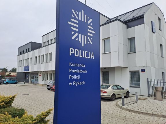 Zdjęcie przedstawia budynek Komendy Powiatowej Policji w Rykach oraz parking przed budynkiem w raz z banerem z logiem policji i nazwa komendy. Budynek ma elewację koloru biało szarego a baner jest niebieski z białymi napisami.