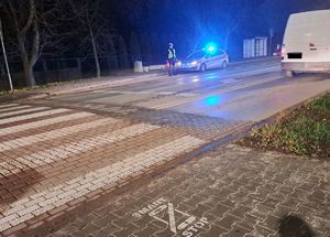 Zdjęcie przedstawia umundurowanego policjanta nadzorującego przejście dla pieszych stojącego obok radiowozu z włączony sygnałami świetlnymi barwy niebieskiej