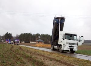 Zdjęcie przedstawia pojazd ciężarowy koloru białego z naczepa typu wywrotka z uniesiona w górze skrzynią ładunkową koloru niebieskiego. Nad pojazdem widoczna jest linia sieci energetycznej. pojazd stoi na drodze asfaltowej a wokół są pola uprawne w oddali widoczne pojazdy służb ratunkowych.