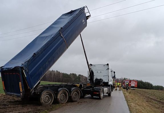 Zdjęcie przedstawia pojazd ciężarowy koloru białego z naczepa typu wywrotka z uniesiona w górze skrzynią ładunkową koloru niebieskiego. Nad pojazdem widoczna jest linia sieci energetycznej. pojazd stoi na drodze asfaltowej a wokół są pola uprawne w oddali widoczne pojazdy służb ratunkowych.