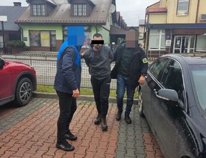 Dwóch policjantów pionu kryminalnego prowadza podejrzanego z założonymi kajdankami. Obok stoją samochody w tle ulica i zabudowa miasta.