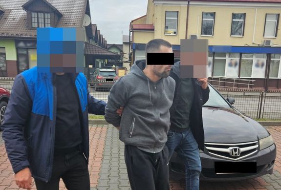 Dwóch policjantów pionu kryminalnego prowadza podejrzanego z założonymi kajdankami. Obok stoją samochody w tle ulica i zabudowa miasta.