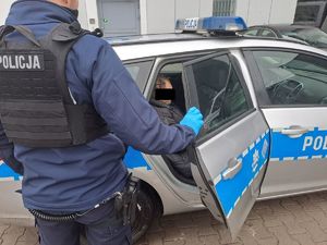 |Męzcz7yzjhna zatrzymany siedzi na tylnej kanapie w radiowozie policyjnym oznakowanym obok stoi policjant odwrócony plecami. Drzwi pojazdu otwarte.