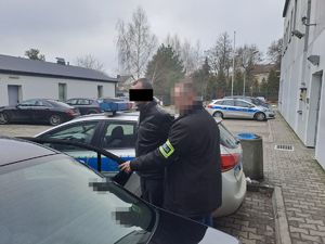 Osoba zatrzymana doprowadzana przez nieumundurowanego policjanta do samochodu na placu komendy policji. |Osoby ubrane w ciemnych ubraniach maja zanonimizowane twarze.