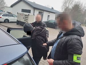 Zatrzymany mężczyzna jest wprowadzany do pojazdu służbowego przez dwóch policjantów kryminalnych w ubraniach cywilnych. Wszyscy mają zanonimizowane twarze. W tle budynki i zaparkowane samochody.