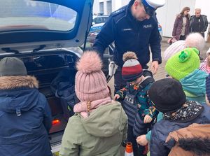 Zdjęcie przedstawiają uczniów,  i nauczycieli szkoły podstawowej w Nowej Rokitni podczas zwiedzania Komendy Powiatowej Policji w Rykach. Dzieci przymierzają kamizelkę kuloodporna i oglądają radiowóz oznakowany i inny sprzęt.