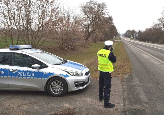 Policjant umundurowany dokonujący pomiaru prędkości na drodze. Obok sto zaparkowany radiowóz oznakowany koloru srebrnego z niebieskimi naklejkami z napisem policja.