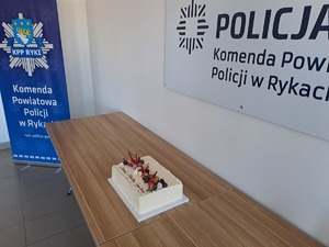 Obchody święta Służby Cywilnej w Komendzie Powiatowej Policji w Rykach - okolicznościowy tort na stole w tle baner z logiem komendy powiatowej policji w rykach