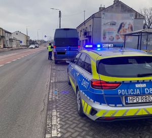 Policjanci ruchu drogowego podczas kontroli pojazdu. Policjant stoi przy pojeździe typu bus koloru niebieskiego oznakowany radiowóz ma włączone sygnały błyskowe niebieski. W tle widoczna droga oraz zabudowania i pojazdy.