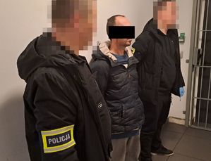 Zatrzymany mężczyzna podczas doprowadzenia w towarzystwie dwóch policjantów nieumundurowanych stojących po obu jego stronach. w Tle wnętrze budynku - korytarz - białe ściany.